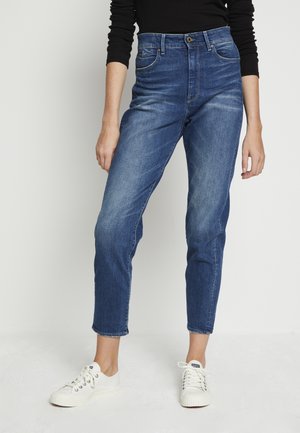 Person, die blaue Jeans mit hoher Taille und schmal zulaufendem Bein sowie weißen Turnschuhen trägt, steht vor einem schlichten hellen Hintergrund.