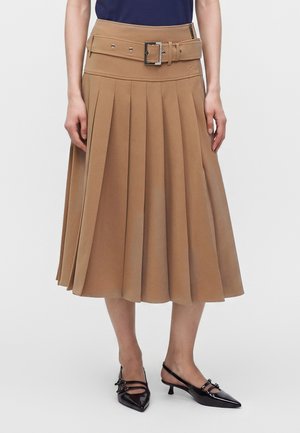 Femme portant une jupe midi plissée beige avec une large ceinture et des escarpins slingback noirs à bout pointu avec de petits nœuds.