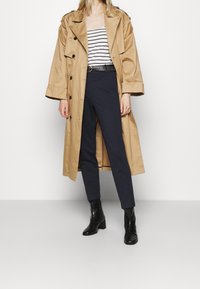 Vrouw in een beige trenchcoat over een gestreept top, marineblauwe tapered broek met een zwarte riem en zwarte hakken enkellaarzen, staande op een grijze vloer.