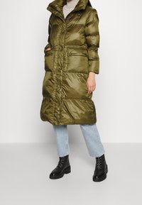 Giacca imbottita verde oliva con colletto alto, dotata di una cerniera frontale, tasche con bottone e una silhouette lunga, indossata sopra jeans chiari e stivali neri.