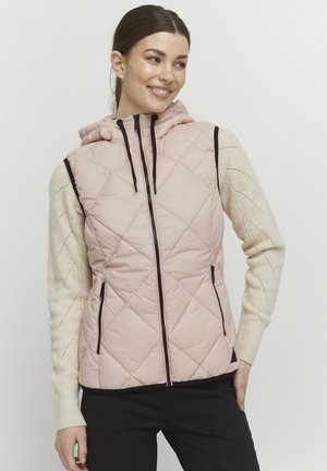 Gilet matelassé rembourré en rose clair avec une capuche, présentant des accents de fermeture éclair noirs et des poches latérales, porté par-dessus un pull couleur crème.