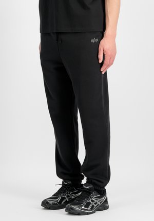 Mann trägt schwarze Jogginghosen mit Logo am Oberschenkel und schwarze Laufschuhe mit weißen Details, steht vor weißem Hintergrund.