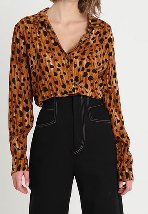 Kobieta w brązowej bluzce z długim rękawem w zwierzęcy wzór, wpuszczonej w spodnie z wysokim stanem w kolorze czarnym z białymi szwami.