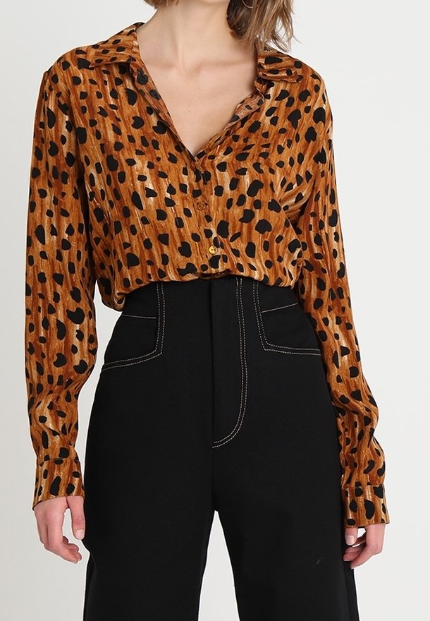Femme portant un chemisier marron à manches longues à imprimé animal, rentré dans un pantalon noir taille haute avec des coutures blanches.