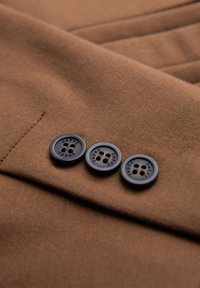 Trois boutons noirs avec le mot "OMBRE" cousu sur une manche en tissu marron d'un vêtement.