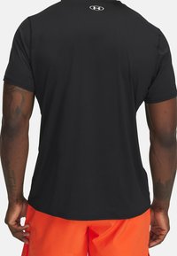 Miehen selkä mustassa Under Armour -lyhythihaisessa paidassa ja kirkkaan oransseissa Shortseissa, jossa näkyy käsivarsitatuointeja ja tumma ihonväri.