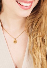 Gouden ketting met ronde hanger met een gestileerde letter "D". Ketting is delicaat en staat tegen een lichtgekleurde top. Haar is golvend.