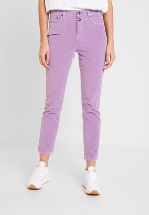 Femme portant un pantalon taille haute, ajusté, de couleur lavande avec une fermeture à double bouton et des baskets blanches, debout devant un fond uni.