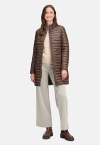 Kvinde iført en brun kviltet lang jakke, beige sweater, ternede vide bukser og brune støvler, står og smiler mod en hvid baggrund.