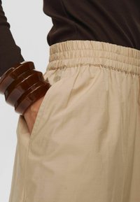 Pantalons beiges à taille élastique avec poches latérales, fabriqués en tissu léger et lisse. Accompagnés d'un bracelet brillant marron au poignet.