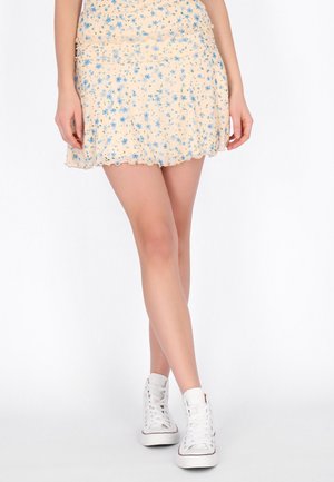 Frau trägt einen hellbeigen Minirock mit blauem Blumenmuster und weißen High-Top-Sneakern, steht mit gekreuzten Beinen vor weißem Hintergrund.