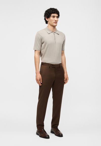 Άνδρας που στέκεται φορώντας μπεζ κοντομάνικη πόλο με φερμουάρ, σκούρο καφέ παντελόνι και σκούρα καφέ επίσημα παπούτσια σε απλό λευκό φόντο.