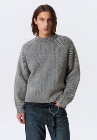Jeune personne avec des cheveux bruns mi-longs portant un pull gris et noir à rayures et un jean sombre avec un grand porte-clés en métal.