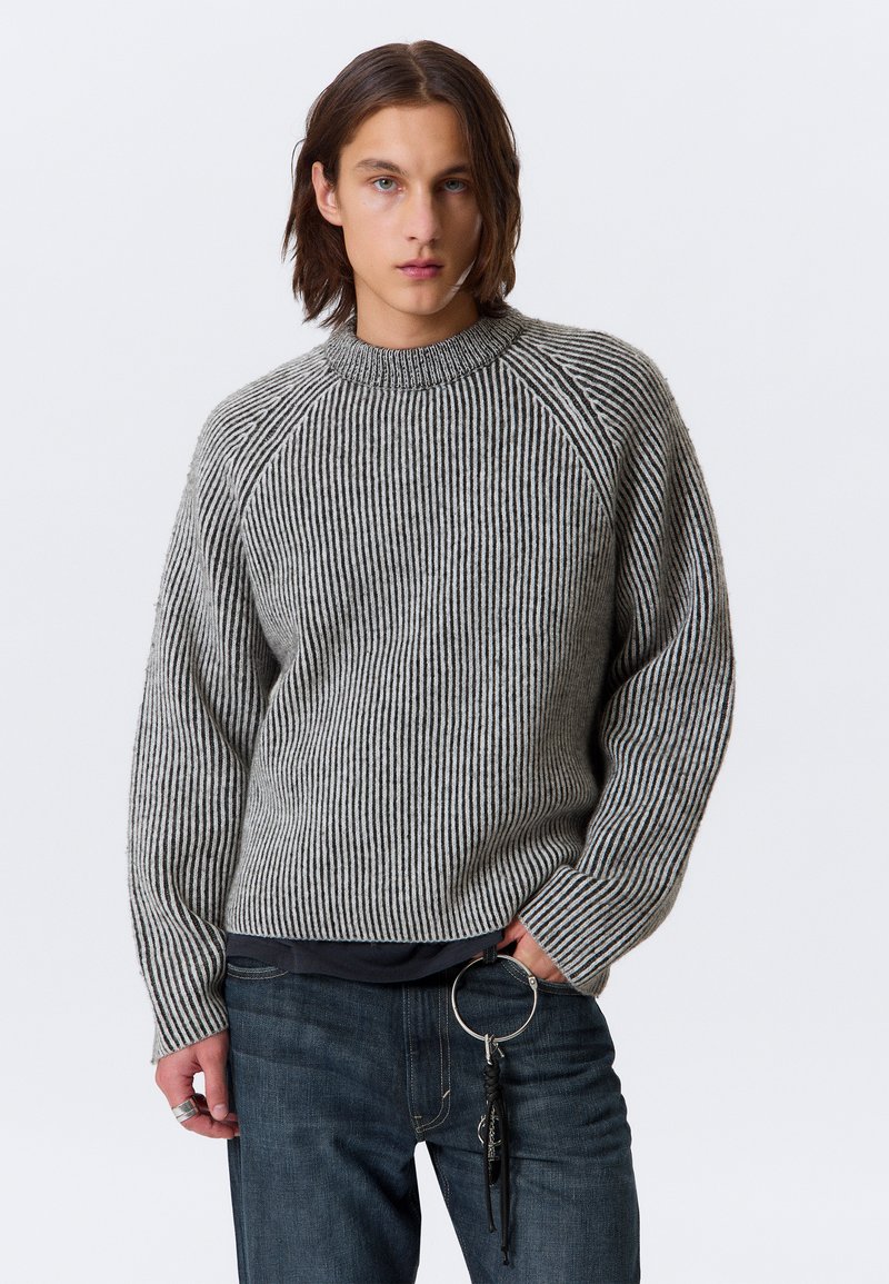 Jeune personne avec des cheveux bruns mi-longs portant un pull gris et noir à rayures et un jean sombre avec un grand porte-clés en métal.