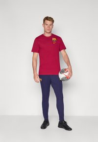 Rød bomulds T-shirt med Barcelona-logo, parret med teksturerede marineblå bukser, holder en sort-hvid mønstret fodbold og har sorte sko på.