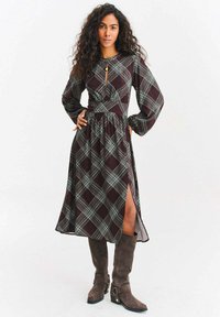 Sukienka w kratę z długim rękawem, z dopasowaną talią i rozcięciem z przodu. Kolory obejmują ciemny bordo i jasnoniebieskie akcenty. Zamszowe kozaki za kolano.