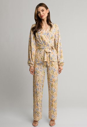 Kobieta ubrane w beżową, kwiecistą bluzkę kopertową z długim rękawem oraz pasujące szerokie spodnie, z odkrytymi palcami na obcasach, stoi na jednolitym tle.