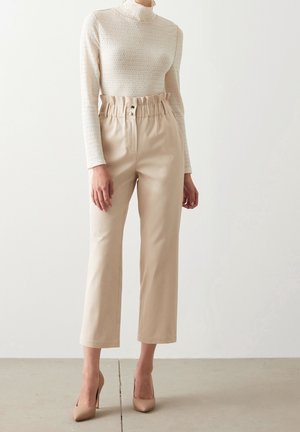 Kobieta ubrana w beżowe spodnie z wysokim stanem typu paperbag, kremową fakturowaną bluzkę z długim rękawem i beżowe szpilki z ostrym noskiem, stojąca na szarej podłodze.