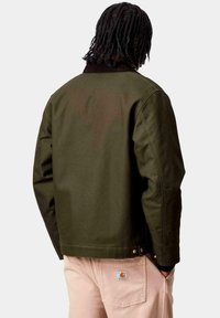 Persona con capelli intrecciati che indossa una giacca verde oliva e pantaloni beige chiaro con un patch del logo Carhartt, voltata di spalle con le mani nelle tasche.
