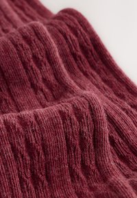 Bordowa dzianina z teksturowanym wzorem warkocza. Miękka, lekko puszysta struktura z naprzemiennymi prążkami i zagłębieniami.