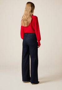Donkere denim jeans met wijd uitlopende pijpen en een rafelige zoom, gecombineerd met een rood gebreide trui met structuur. De foto toont de achterkant van de outfit.