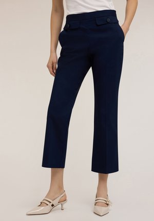 Marineblaue Cropped-Hosen mit zwei Fronttaschen und flachem Vorderteil, ausgestattet mit dekorativen Knöpfen. Getragen mit beigen Slingback-Absätzen.