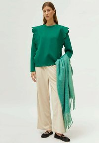Sudadera verde con detalles en forma de concha, combinada con pantalones de pierna ancha a rayas crema y una bufanda de gran tamaño en color teal, llevada con zapatos planos negros.