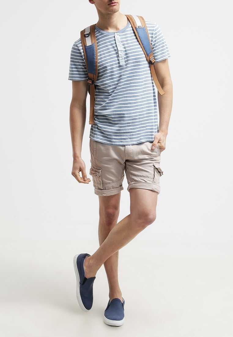 Chemise à manches courtes rayée bleu et blanc, short cargo beige, chaussures nautiques bleu marine et un sac à dos marron avec des accents bleus.