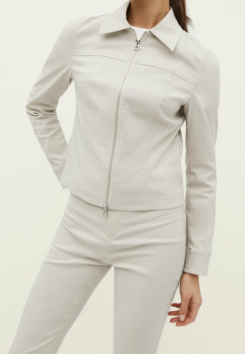 Lysegrå lynlåstrøje med krave, skræddersyet pasform og glat stof. Har syninger over brystet og lange ærmer.