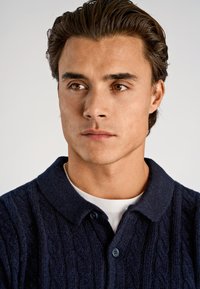 Marineblaues, kabelgestricktes Poloshirt mit strukturierter Oberfläche, versehen mit klassischem Kragen und zwei dunklen Knöpfen, über einem weißen Hemd getragen.