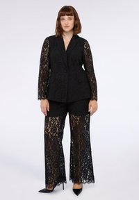 Completo in pizzo nero con blazer a maniche lunghe e pantaloni a gamba larga. Il tessuto presenta un intricato motivo floreale ed è semitrasparente.