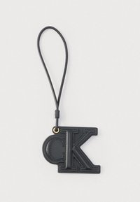 Porte-clés en cuir noir avec les lettres embossées "C" et "K" attaché à une sangle en boucle sur un fond blanc.