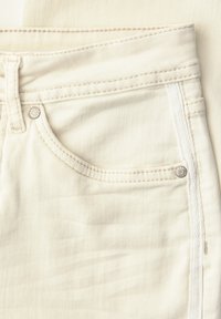 Primer plano de un bolsillo de jeans de mezclilla color crema, con costuras, presilla para cinturón y remaches metálicos en el borde del bolsillo.