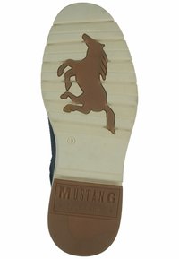 Semelle en caoutchouc avec une base blanche et un logo de cheval marron en relief. Présente des rainures horizontales pour la traction et la marque "MUSTANG True Denim".