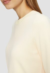Jasna kremowa sweter z okrągłym dekoltem i teksturowanym szwem na ramionach. Materiał wydaje się miękki i ma dopasowany krój.