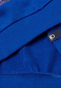 Modrý svetr ze tkaniny s pružnými manžetami a pasem. Obsahuje strukturovanou vnitřní vrstvu a malou nášivku s logem na boku.