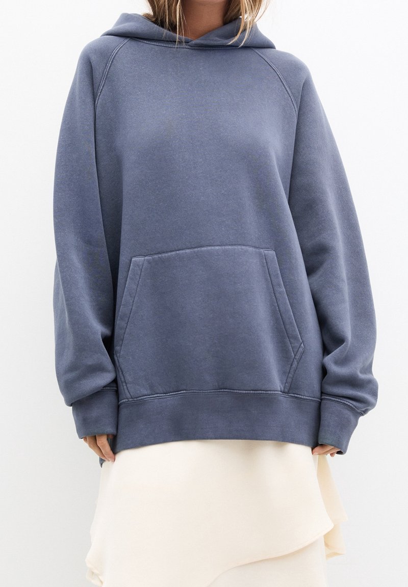 Femme portant un sweat à capuche bleu ample avec une poche frontale et une jupe crème à couches sur fond uni.