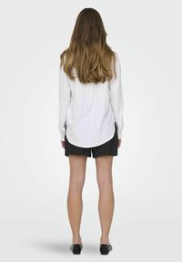 Camicia bianca a maniche lunghe con una vestibilità morbida, abbinata a pantaloni corti neri. La camicia ha un orlo curvo e la figura indossa scarpe flat nere.