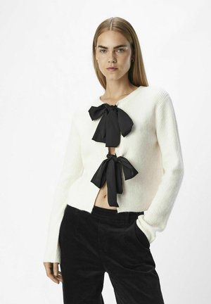 Vit ribbad cardigan med två svarta rosetter framtill, med en något beskuren längd, i kombination med svarta manchesterbyxor.