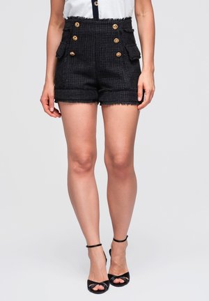Donna che indossa pantaloncini neri in tweed a vita alta con bottoni dorati e sandali neri con tacco alto e cinturino alla caviglia, in piedi su uno sfondo semplice.