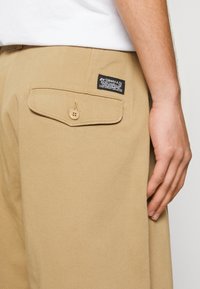Pantaloni beige con cuciture visibili, tasca a patta sul retro e un'etichetta nera con scritte bianche. Il tessuto sembra liscio e resistente.
