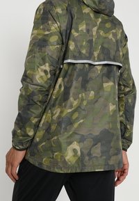 Man met een groen en bruin camouflage Nike-jack met capuchon, reflecterende strip op de rug en zwarte broek, met zijn rug naar de camera gekeerd.