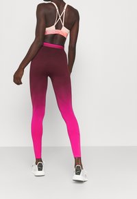 Kobieta stoi na jednolitym tle, ubrana w różowo-fioletowe legginsy w ombre, brzoskwiniowy stanik sportowy z krzyżującymi się ramiączkami i białe sneakersy.