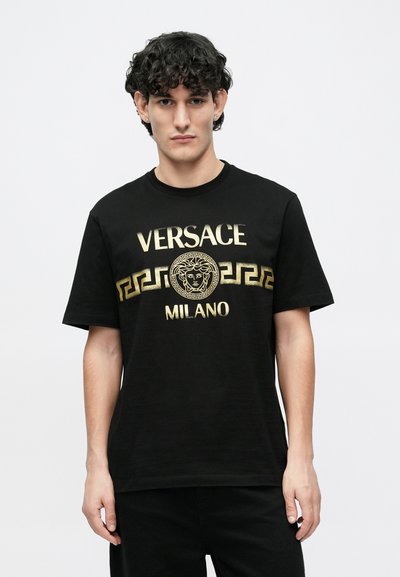 Černé tričko s krátkými rukávy, s textem „VERSACE MILANO“ ve zlaté barvě a centrálním emblémem Medusy s řeckým klíčovým vzorem pod ním.