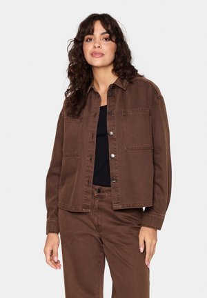 Braune Denimjacke mit Kragen, zwei Vordertaschen und Druckknopfverschluss. Über einem schwarzen Oberteil getragen, hat sie lange Ärmel.
