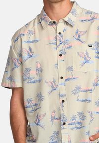 Camisa de manga corta beige con estampado de aves tropicales azules y rosas y palmeras. Presenta un bolsillo en el pecho y botones marrones.