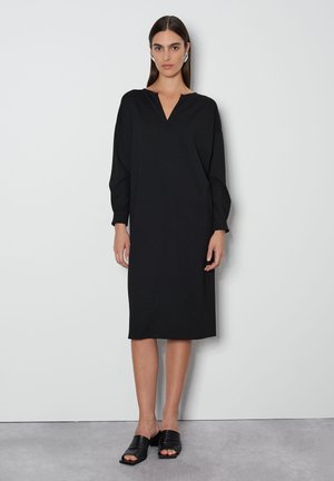Schwarzes langärmliges Kleid mit V-Ausschnitt, gerader Silhouette, knielang, aus weichem Stoff. Getragen mit schwarzen offenen Mules.