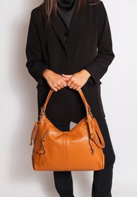 Donna in giacca nera che tiene una grande borsa in pelle beige con hardware dorato e portachiavi su sfondo bianco.