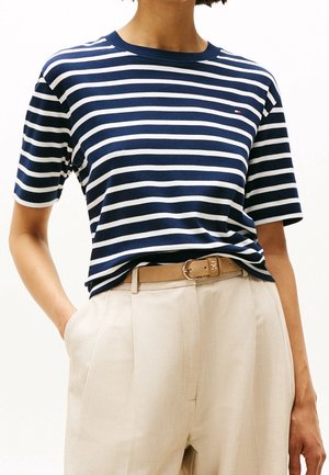 Person iført navyblå og hvidstribet kortærmet skjorte stoppet ned i beige højtaljede bukser med lysebrunt bælte.