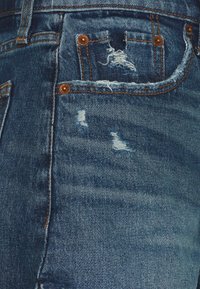 Jean en denim bleu avec des détails usés, des bords effilochés, des rivets en cuivre et des coutures contrastantes marron, le tout sur un tissu à texture douce.